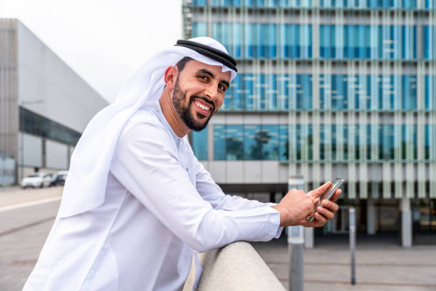 A smiling man in traditional attire leans on a railing with a smartphone, symbolizing business readiness in Ajman.